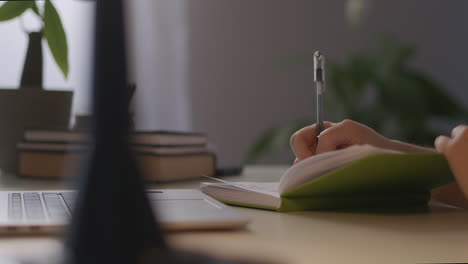 Una-Estudiante-Está-Escribiendo-Notas-En-Un-Cuaderno-Con-Un-Bolígrafo,-Primer-Plano-De-Una-Mesa-De-Trabajo-Con-Computadora-Portátil,-Aprendizaje-Electrónico-Y-Desarrollo-Profesional-De-Educación-En-Línea